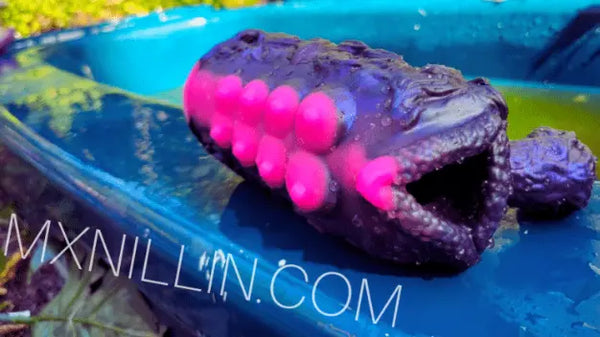 Outdoor photo of a purple fantasy stroker with bright pink suction-cup textures, resting on a blue surface with the watermark ‘MXNILLIN.COM’.