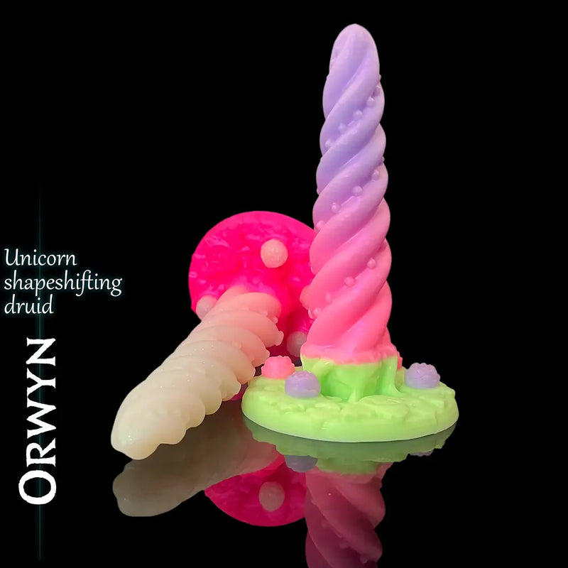 Orwyn Unicorn Shape-Shifting Druid silikonowe dildo fantasy w pastelowy różowy i lavender z zielony rzeźbiony podstawa, prezentowane na błyszczący czarny odbijający światło tło z oznaczeniem nazwy produktu.