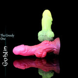 Two "The Greedy One" Goblin fantasy toys stacked against a black background. One is a luminous pink and the other is a luminous green, showcasing two available custom colors.