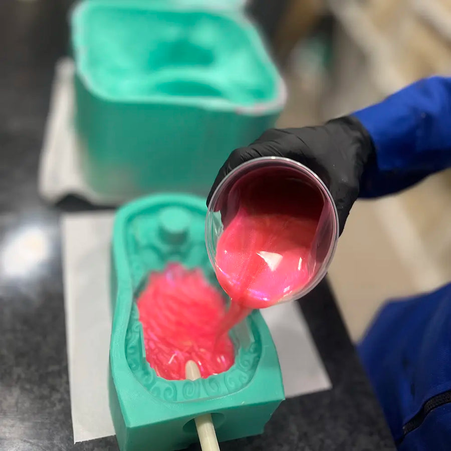 Close-up of a production process showing a person in blue coveralls and black gloves pouring shimmery pink liquid silicone into a turquoise-colored custom mould for a pleasure product in a laboratory setting.